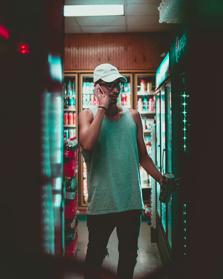 man using smartphone standing in front on beverage cooler inside store, man holding drinks can while putting phone on ear inside store, HD wallpaper