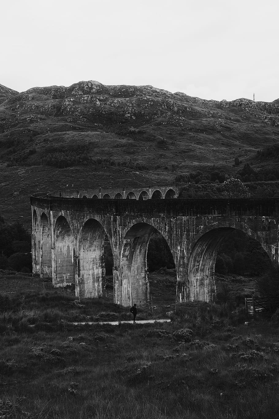 Glenfinnan Viaduct in Black and White, grayscale photo of man standing under concrete building near hills