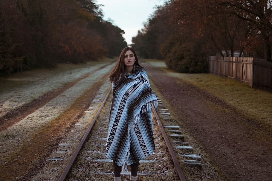 woman with blue and black striped scarf standing on brown train railway during daytime, woman standing on train rail surrounded by trees during daytime, HD wallpaper