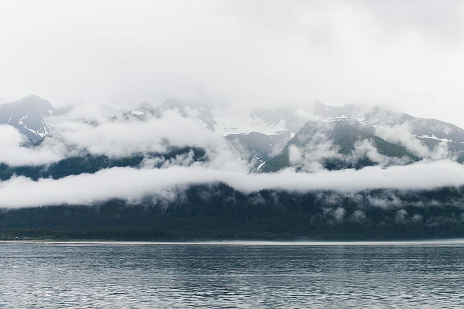cloudy sky covered mountain taken at daytime, gray mountain covered by clouds across body of water