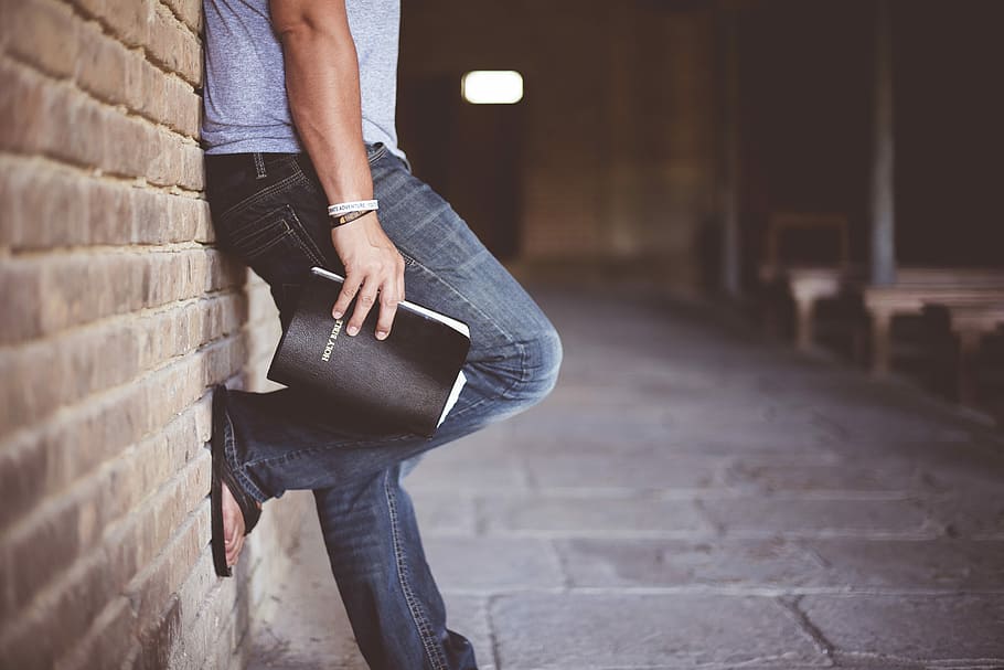 man holding Holy Bible leaning on bricked wall, person leaning on wall