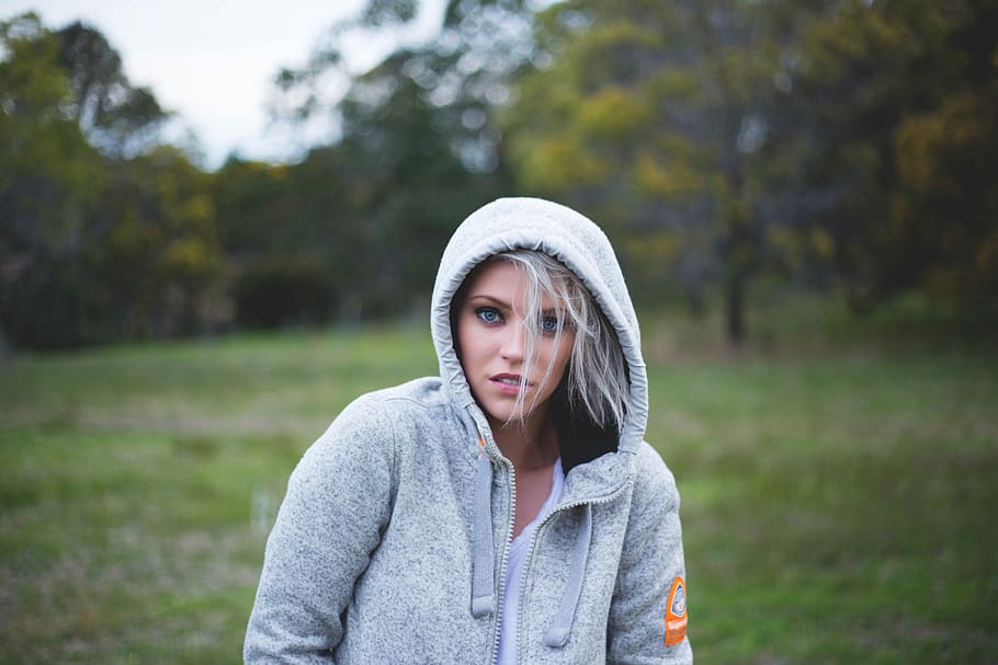 tilt-shift lens photography of woman on a green grass, selective focus photography of woman wearing grey hooded zip-up jacket near tall trees during daytime