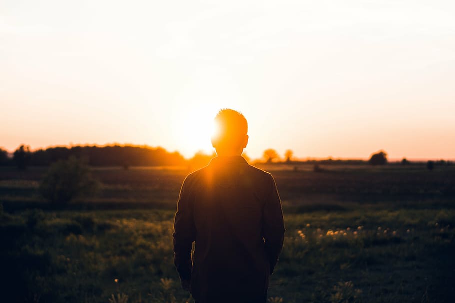 silhouette of man standing on grass field facing mountain range under golden hour HD wallpaper
