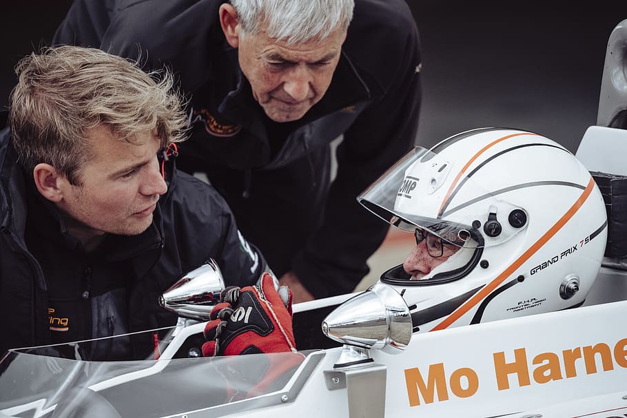 man in black jacket talking to the a wearing white full-face helmet during daytime, selective focus photography of two men standing beside man in white F1 race car at daytime