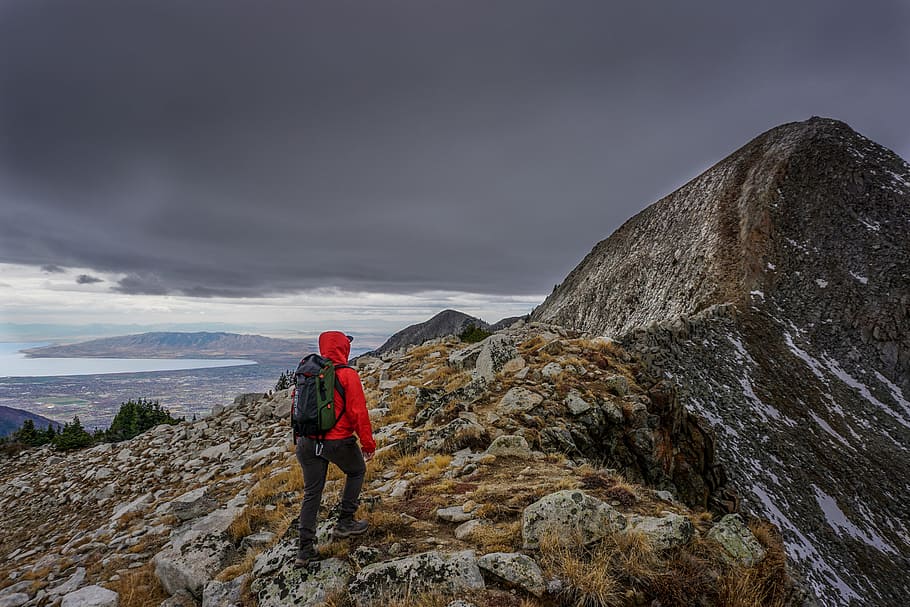 person wearing red hoodie standing on mountain near peak under grey cloudy sky HD wallpaper