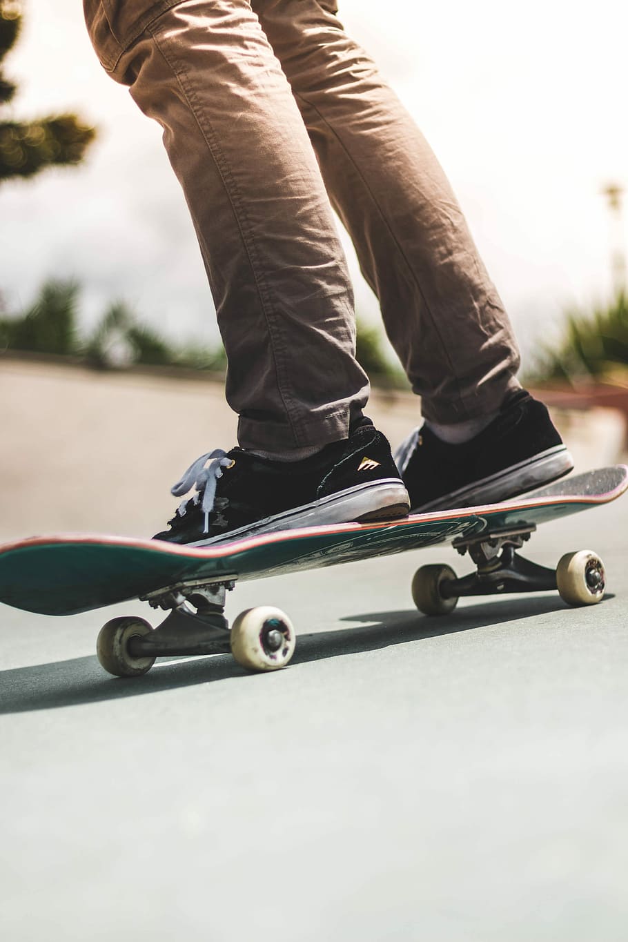person wearing pair of black-and-white Easton low-top sneakers and brown pants while riding on brown and black skateboard on gray concrete road during daytime