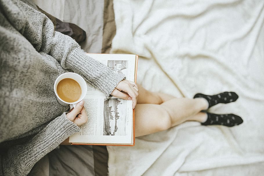 woman holding a cup of coffee at right hand and reading book on her lap while holding it open with her left hand in a well-lit room, woman drinking coffee, HD wallpaper