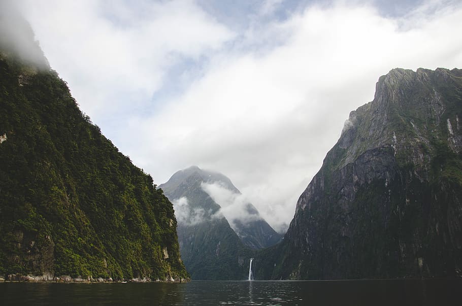 body of water between mountains, calm river near waterfalls and mountains with fogs at daytime HD wallpaper