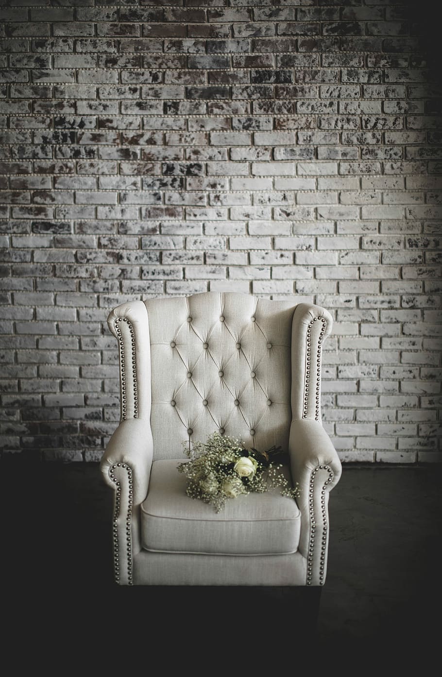 white flowers on top of white armchair, red rose flower bouquet on tufted white fabric armchair in front of gray concrete brick wall