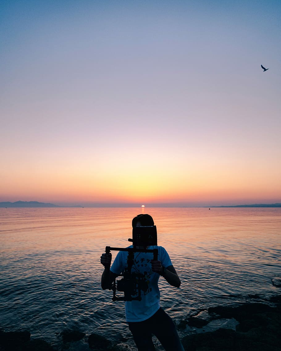 man near body of water, man holding camera stabilizer on seashore during sunset HD wallpaper