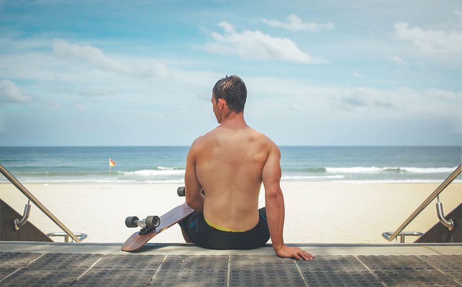 man sitting on staircase holding brown cruiser board near beach during daytime HD wallpaper