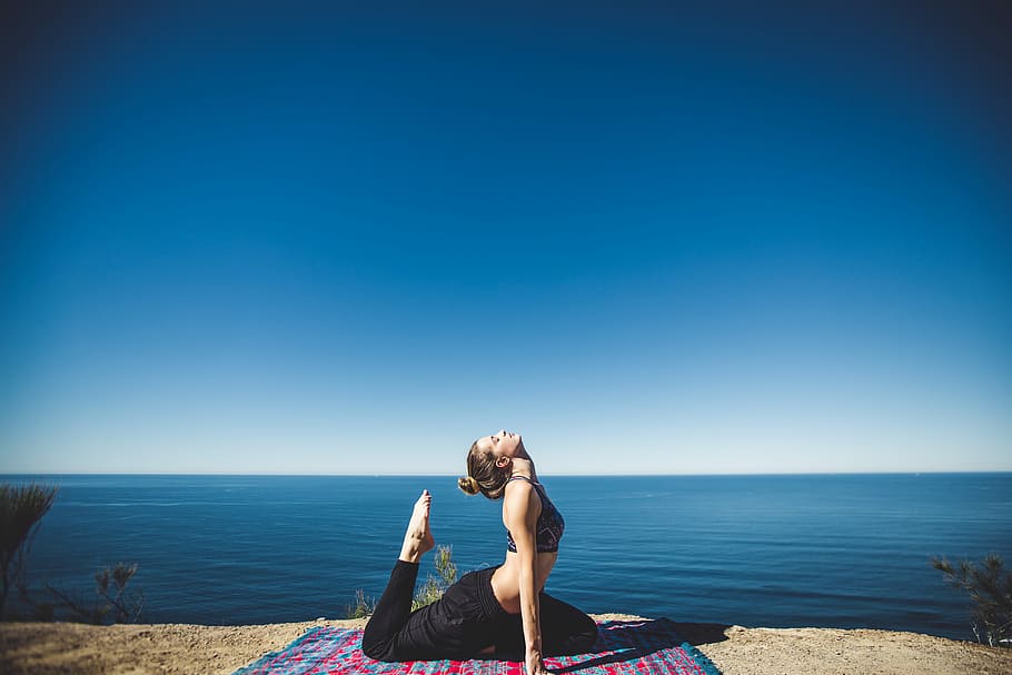 woman wearing black top and pants stretching body on mat, woman wearing black doing yoga beside sea under blue sky
