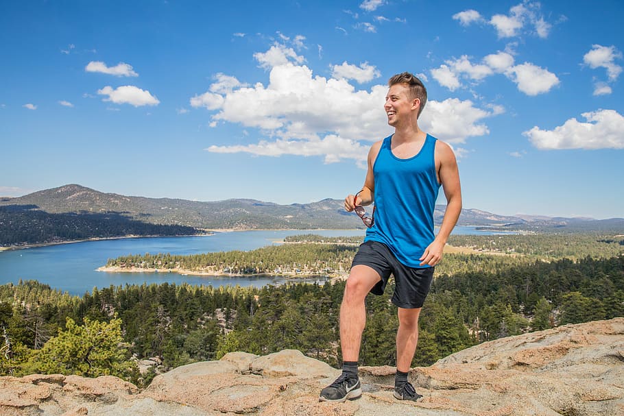 man wearing blue tank top and black shorts standing on rock cliff, man standing on hill with body of water in the background HD wallpaper
