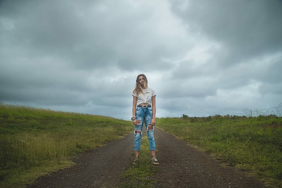 woman standing on dirt road under cloudy sky during daytime, woman standing in middle of road between grass field HD wallpaper
