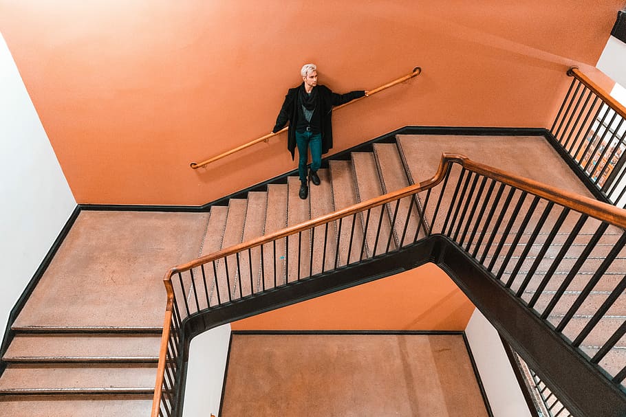 person standing on staircase, person standing and leaning on orange painted wall holding brown handrail on stairway HD wallpaper