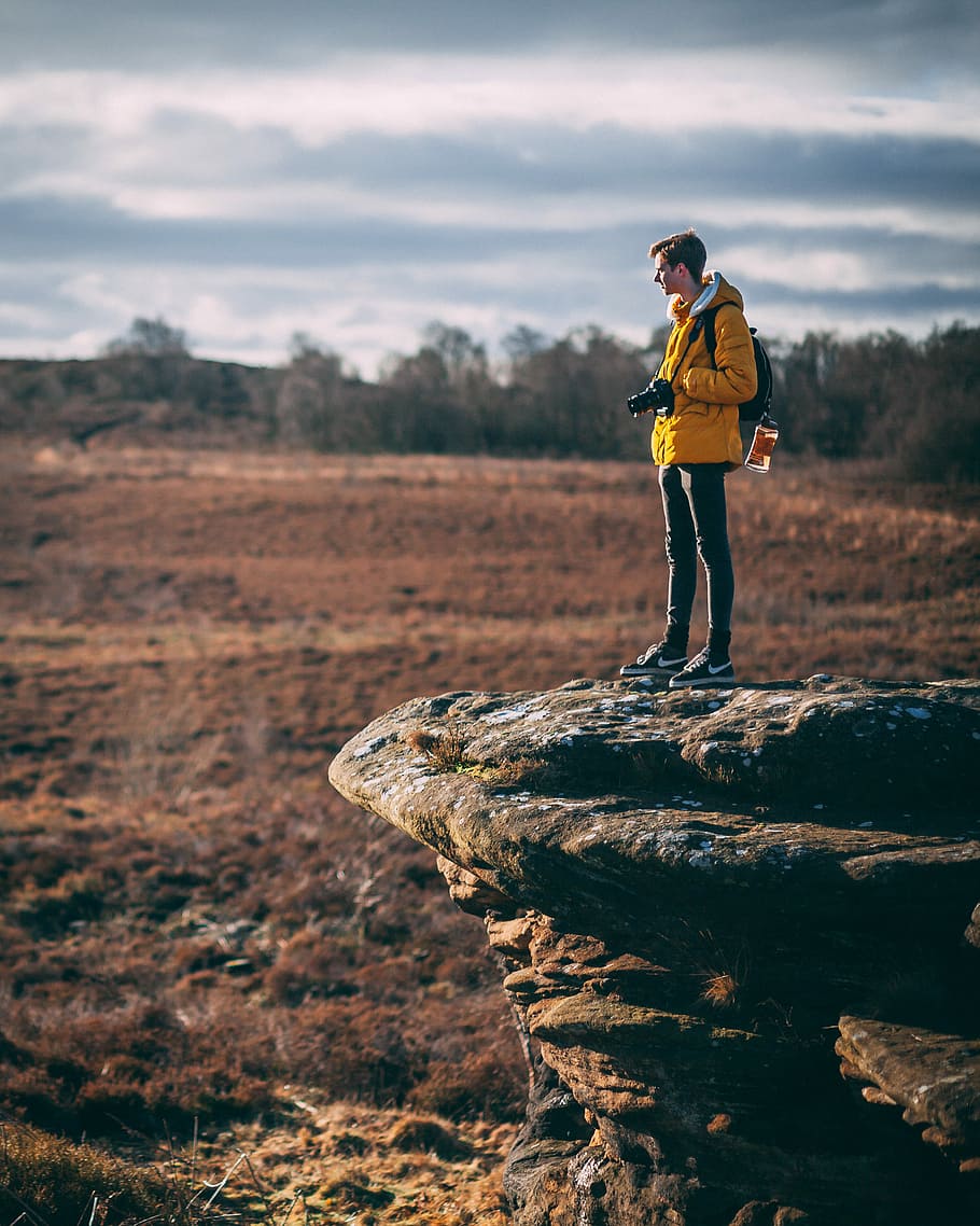 man standing at cliff holding his camera, man standing on gray rock near green leafed trees HD wallpaper