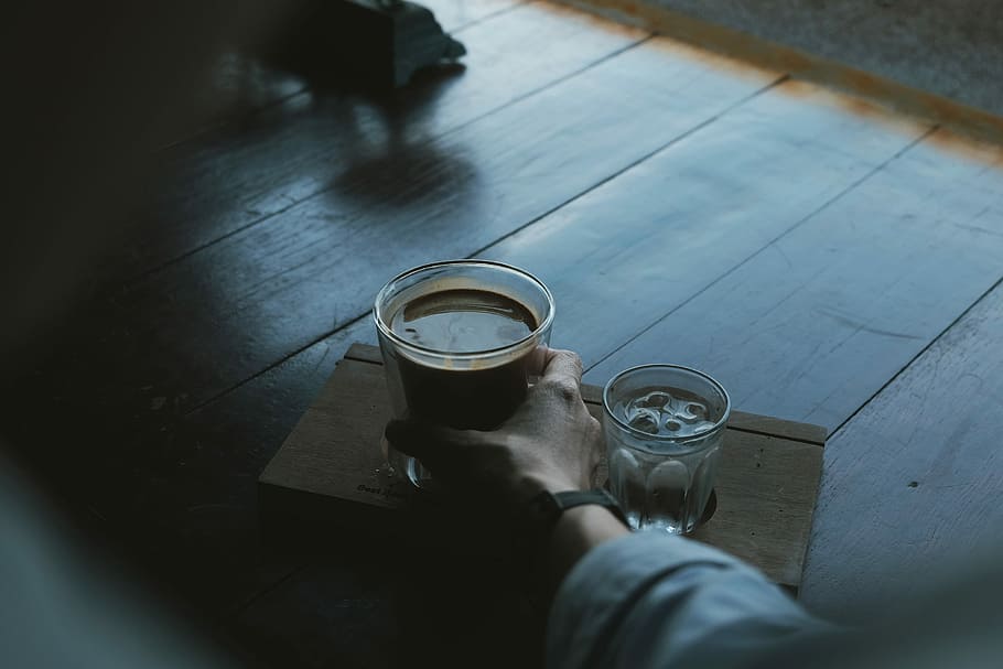 person holding brown liquid filled clear drinking glass beside clear liquid filled glass with ice cubes on brown wooden tray, person holding clear glass mug HD wallpaper