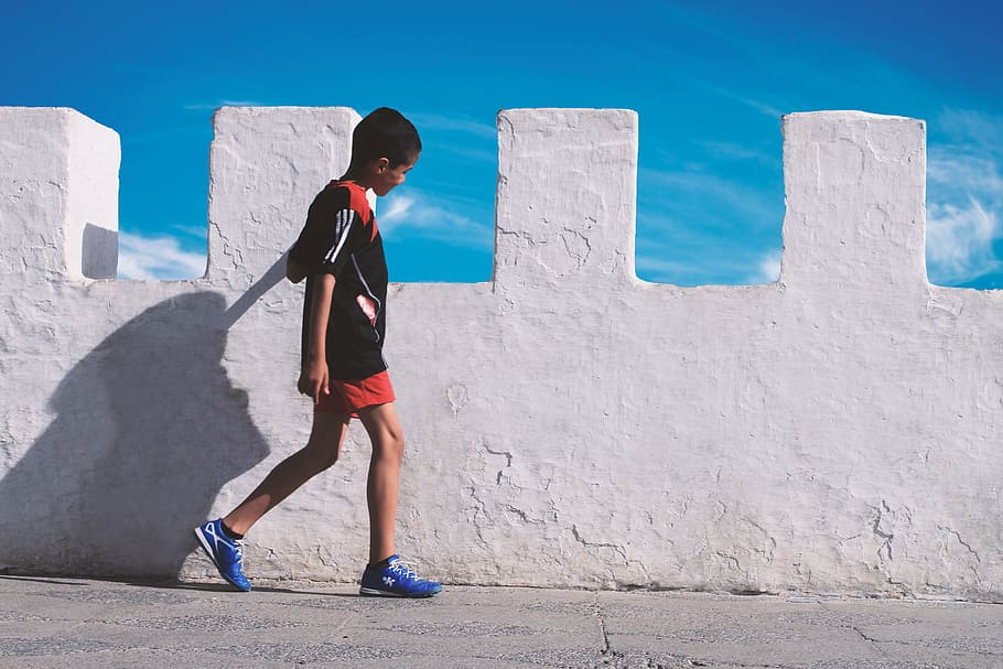 boy walking beside white concrete wall, boy walking neat concrete wall during daytime HD wallpaper