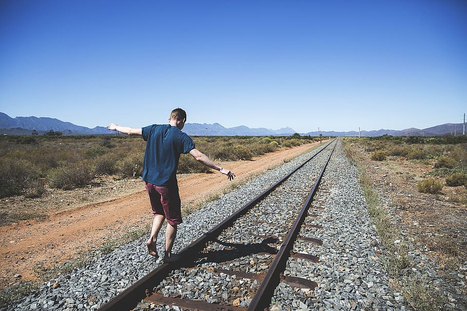 person walking on brown steel train rail outdoor during daytime, man balancing on train railings under blue sky