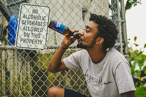 man drinking beverage in amber glass bottle beside gray wire link fence at daytime, photography of man in gray shirt drinking beer during time HD wallpaper