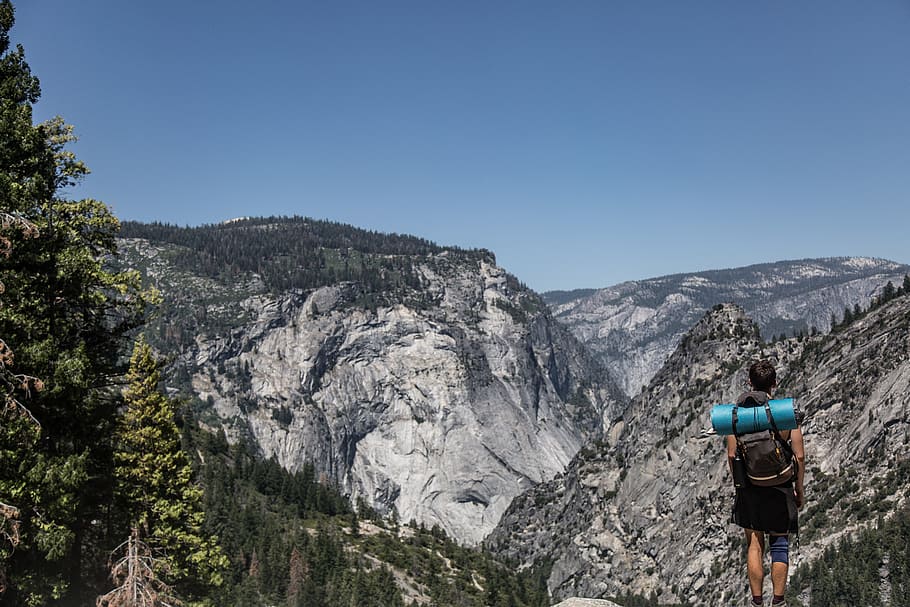 man standing on cliff, man standing on cliff facing mountain landscape during daytime HD wallpaper