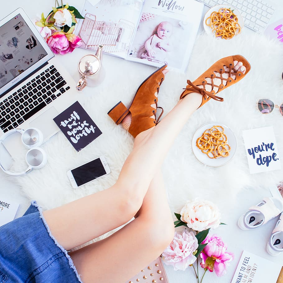 woman wearing brown open-toe chunky sandals sitting beside white smartphone, headphones and laptop, woman sitting on rug surrounded with laptop, flowers, art materials, and shoes flatlay photography