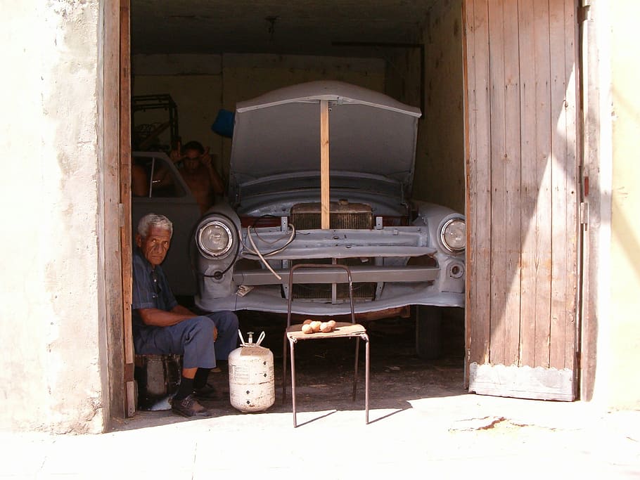 man sitting on stool near white gas tank and classic blue car body shell in garage during daytime HD wallpaper