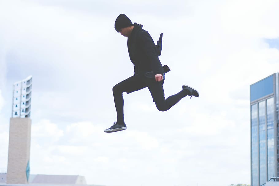 photography of man wearing black beanie, black top, black pants, and black low top sneakers jumping under gray sky HD wallpaper