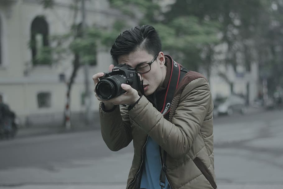 shallow focus photography of man taking photograph holding his camera, man wearing brown zip-up bubble jacket holding DSLR camera taking photo near on road near building during daytime