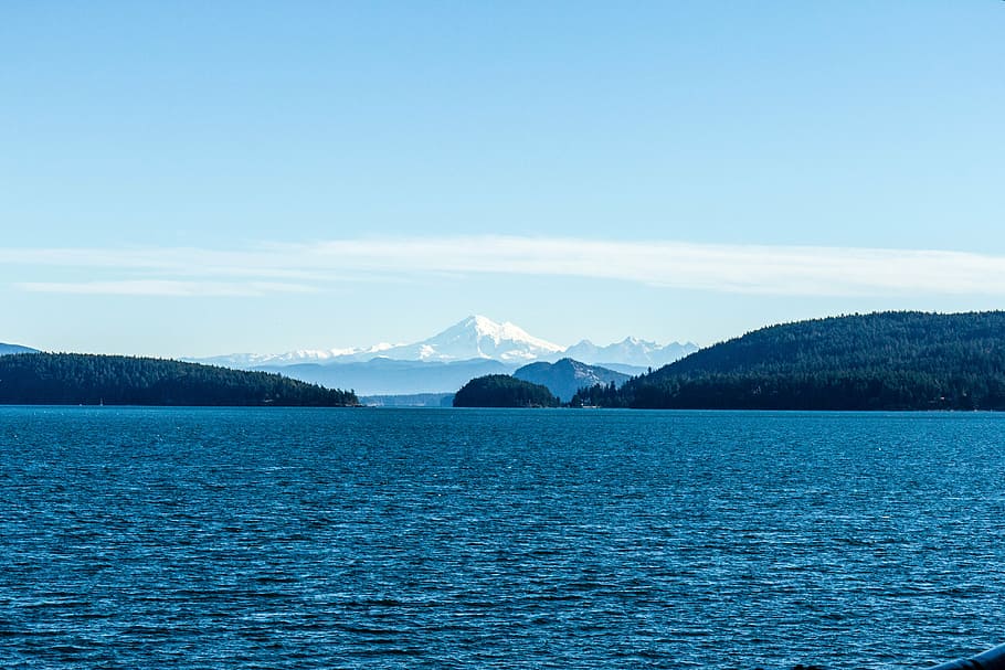 calm body of water overlooking mountain under blue sky at daytime HD wallpaper