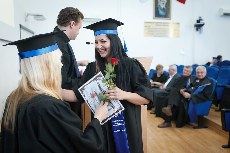 people standing and sitting inside room wearing academic dress and woman holding certificate and red rose smiling