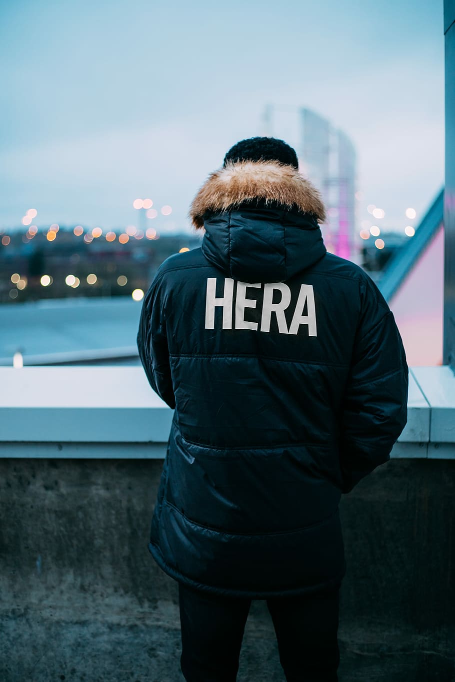 person wearing black and brown parka jacket with Hera-printed text standing facing the cities, person wearing black Hera coat in front of building, HD wallpaper
