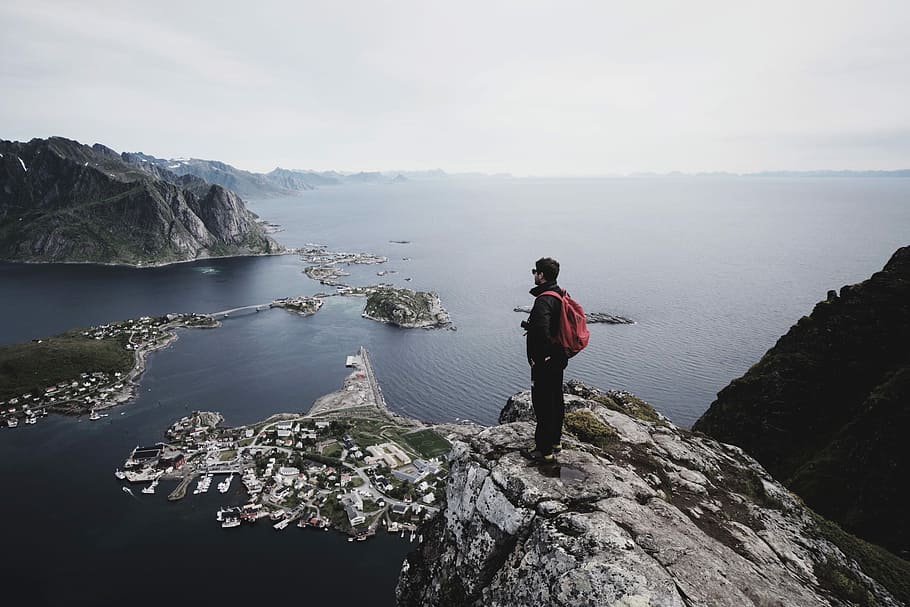 man standing on top of a cliff facing cityscape by water, man standing on cliff during daytime HD wallpaper