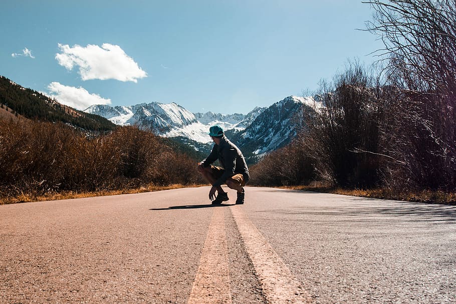 person squatting in the middle of road near snow-capped mountain at daytime, man sitting on road during daytime HD wallpaper
