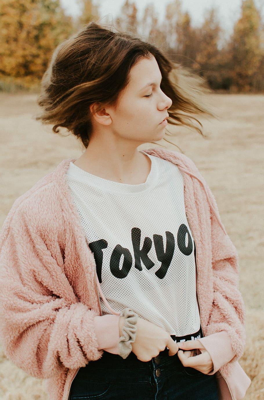 girl wearing pink jacket holding her shirt, selective focus photography of women's white Tokyo-print mesh crew-neck shirt and pink fleece cardigan standing on grass field at daytime, HD wallpaper