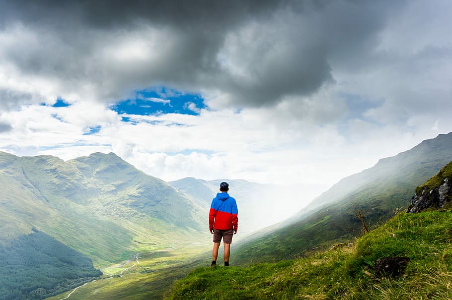 photography of man standing on green grass, man standing on green mountain under cloudy sky during daytime HD wallpaper