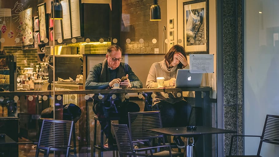 man holding smartphone sitting beside woman in front of silver MacBook in restaurant, man wearing grey coat sitting on chair beside woman while using MacBook