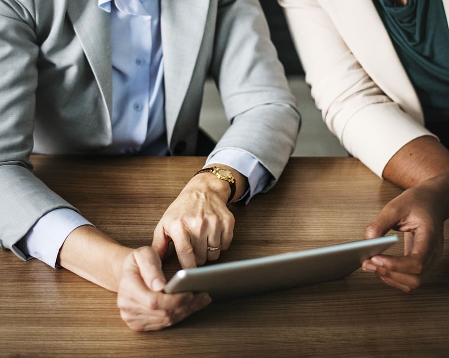 two persons sitting side by side across table and looking at a tablet computer in a well-lit room HD wallpaper