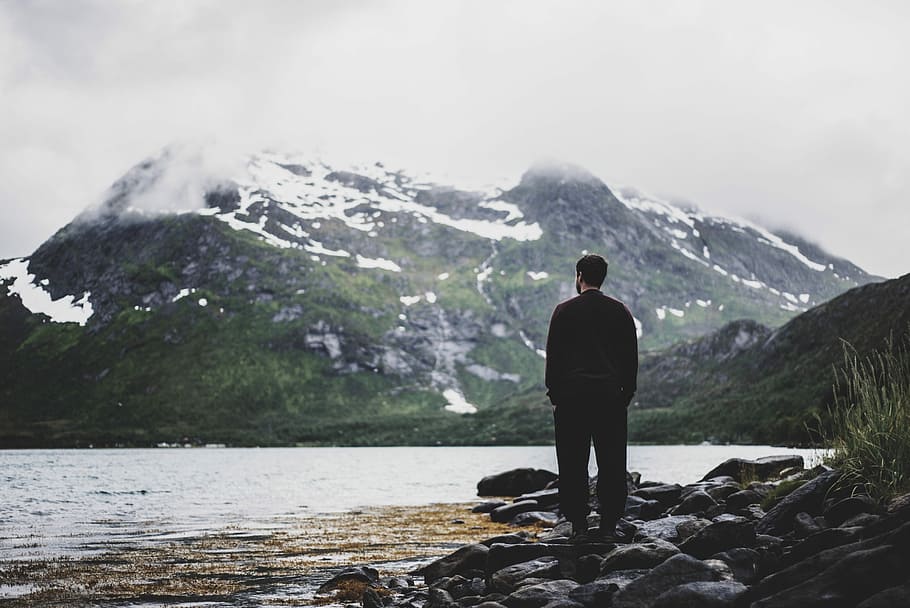 photo of man standing near seashore facing mountain covered with snow, man standing on stone beside the seshore HD wallpaper