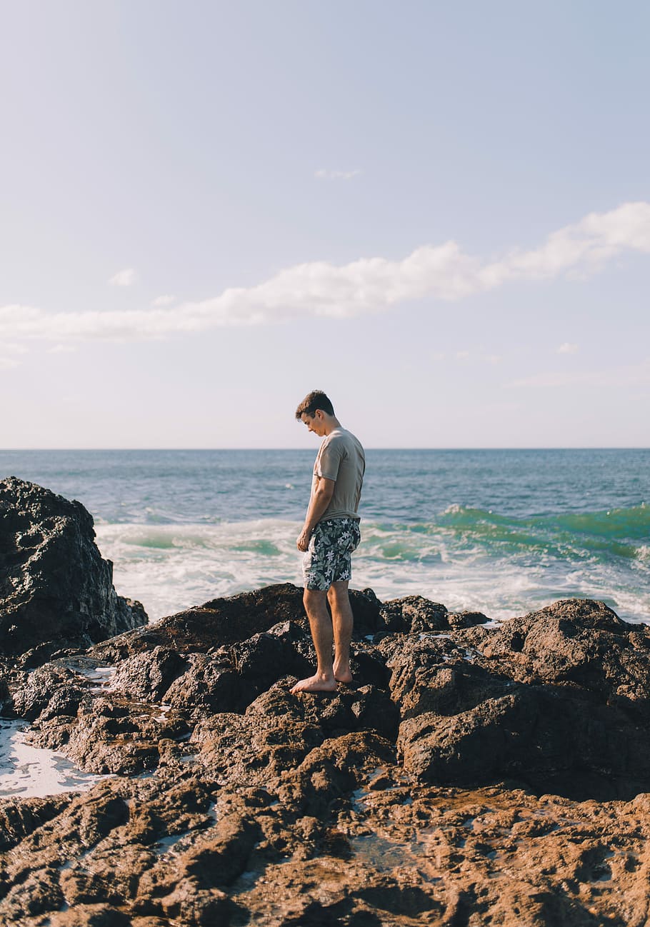 man standing on rock island beside beach, man standing near shore HD wallpaper
