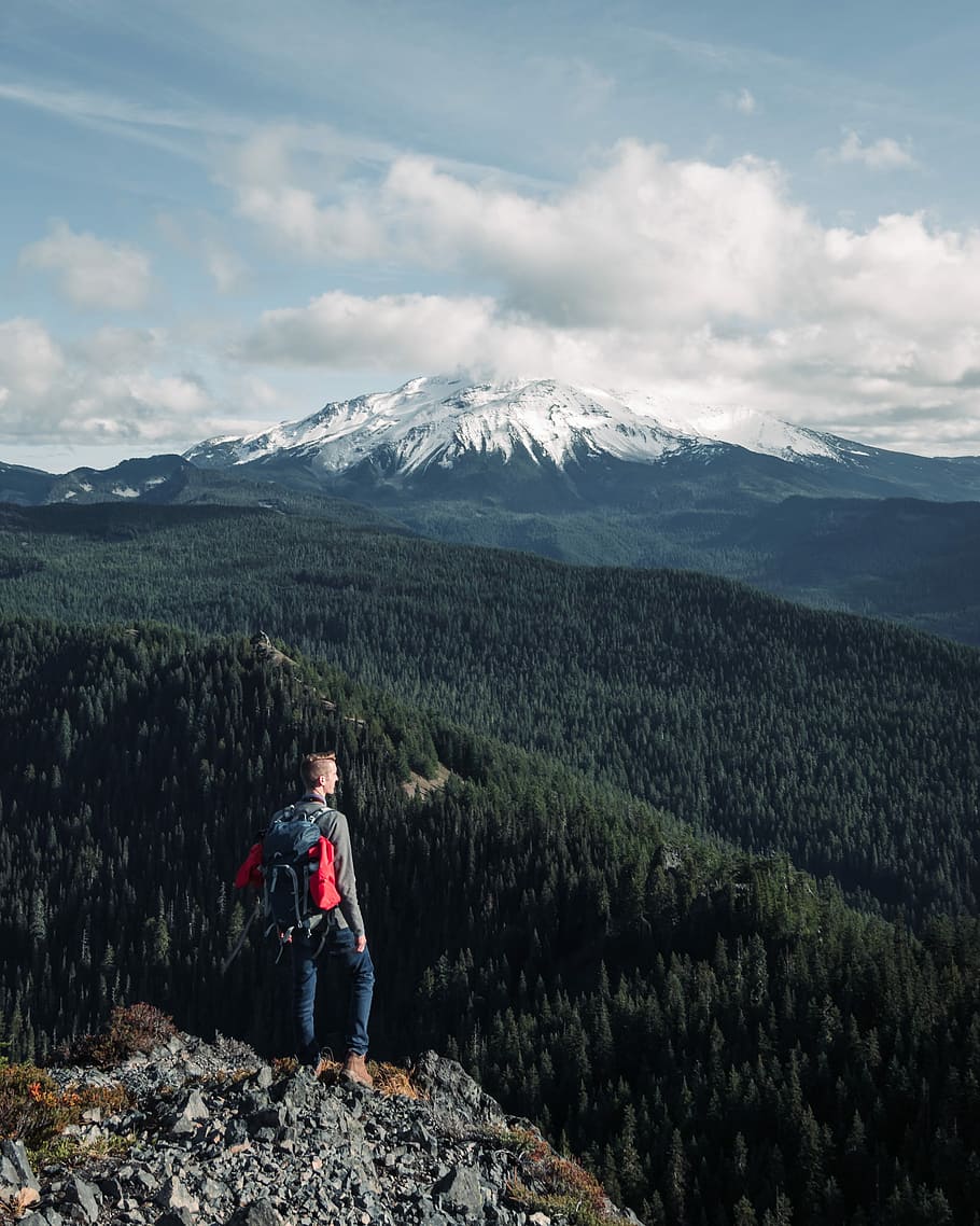 man standing on peak of mountain under cloudy sky, shallow focus photography of man standing on cliff HD wallpaper