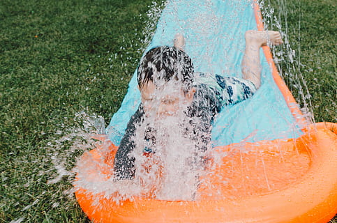 child sliding on blue and orange slippery pad with water splash at daytime, boy on orange and blue inflatable slide splashing water during daytime photo HD wallpaper
