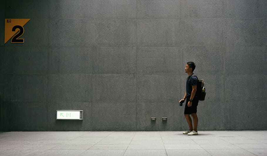 man in black top carrying backpack standing near wall, man in black shirt standing near gray wall