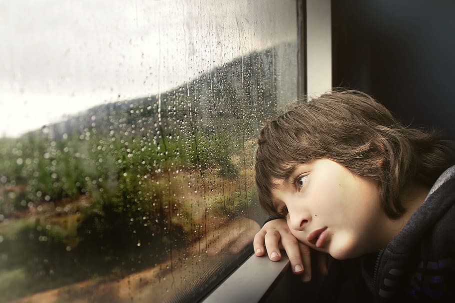 boy resting cheek on hand while looking outside window, person