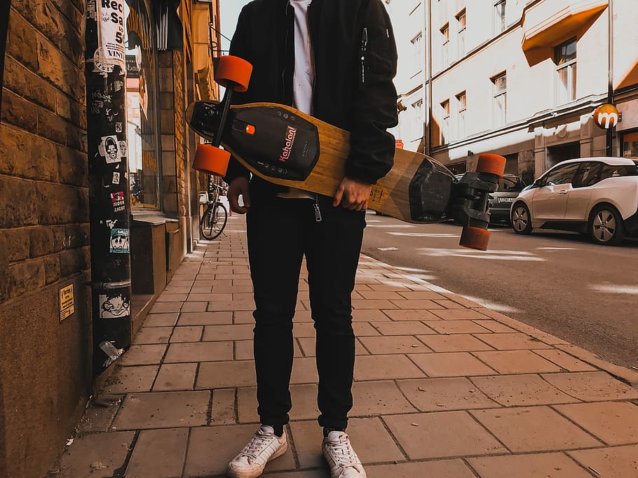 person holding skateboard near building, man wearing black jacket and black pants carrying brown longboard