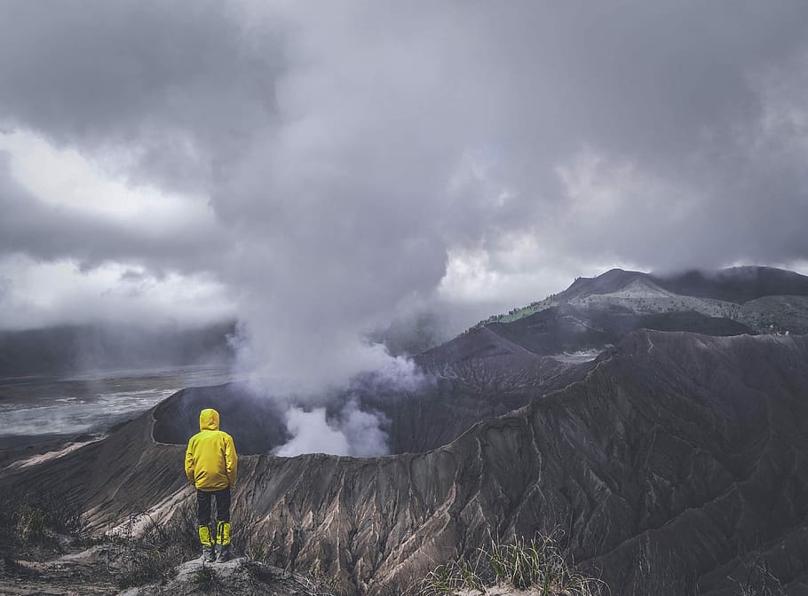 person wearing yellow jacket standing on cliff, person in yellow jacket facing volcano HD wallpaper