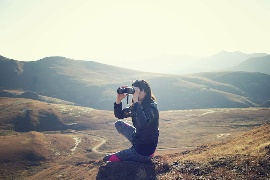 woman sitting on rock while using binoculars during daytime HD wallpaper