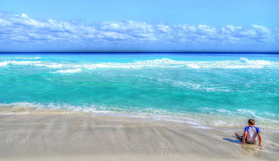man wearing white and blue raglan shirt sitting in seashore facing the blue sea during daytime HD wallpaper