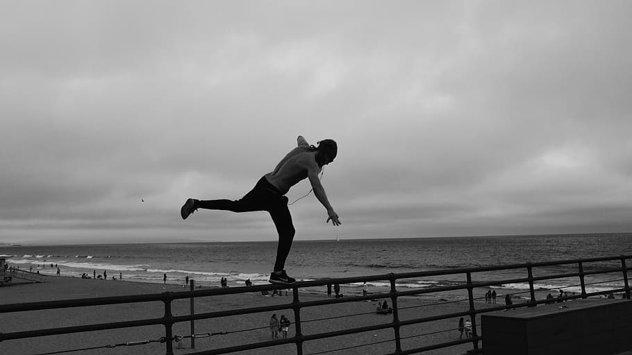 man standing on metal railing overlooking beach shoreline under gray cloudy skies at daytime, man balancing on railing near the beach HD wallpaper