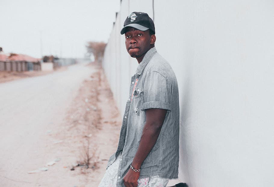 man in gray shirt and black cap leaning on white wall, man stands beside white painted wall outdoor during day HD wallpaper